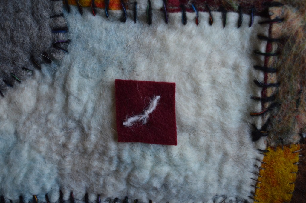 Details of Textured felt artwork with earthy patchwork in brown, yellow, and grey, featuring a central fuzzy red starburst and embroidered white crosses, reflecting layered memory.