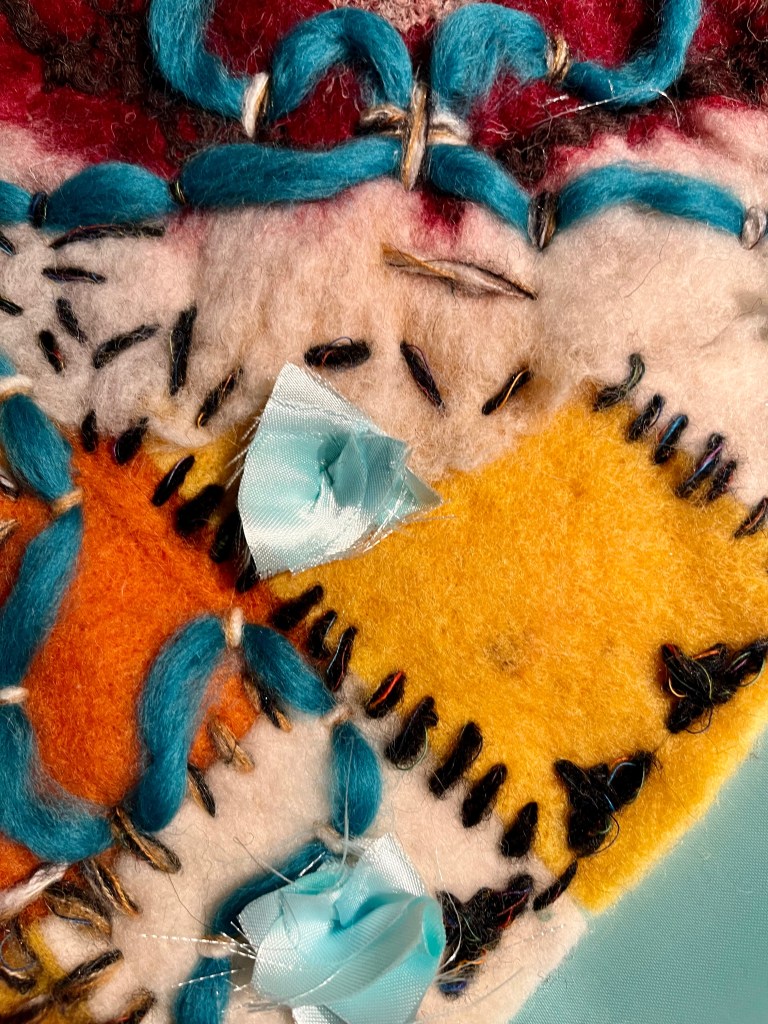 Handmade felt collage with stitched patchwork squares in ochre, white, and grey, centered around a deep red motif, adorned with turquoise merino wool and small blue textile accents.