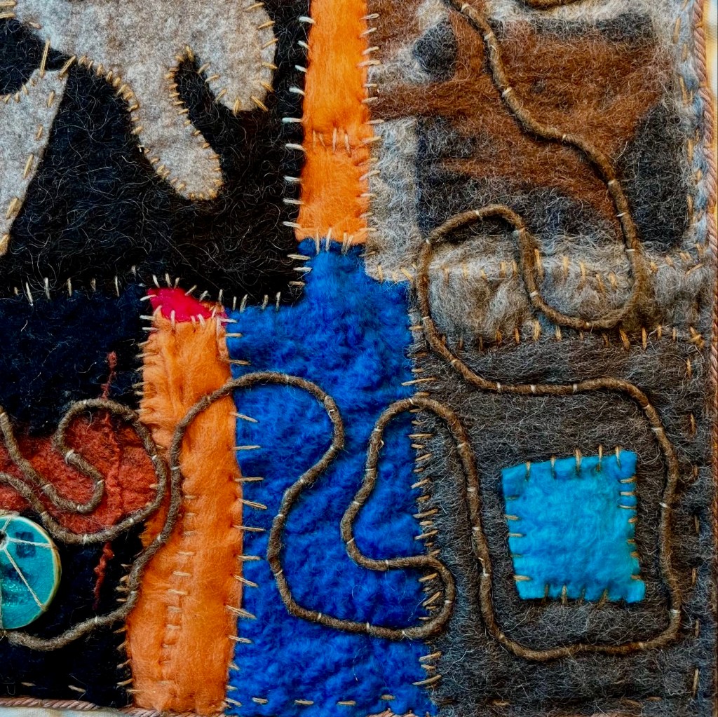 Square felt artwork with a bold symmetrical face-like motif in cream and black, surrounded by winding yarn lines, bright accents, and stitched circular emblems — blending tribal rhythm with mystical geometry.