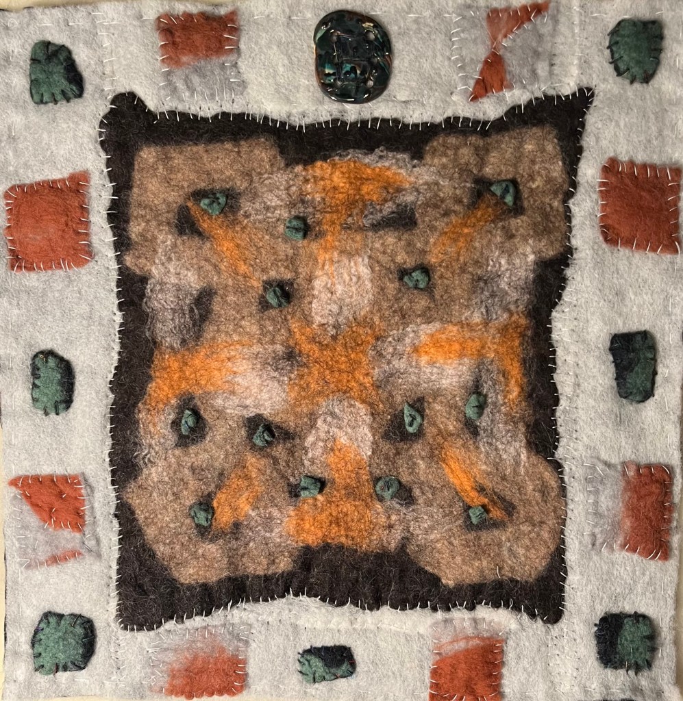 Earthy-toned felt collage with a rust and brown patterned center surrounded by stitched pale fabric squares and a dark green ceramic element at the top — blending textile warmth with sculptural accent.