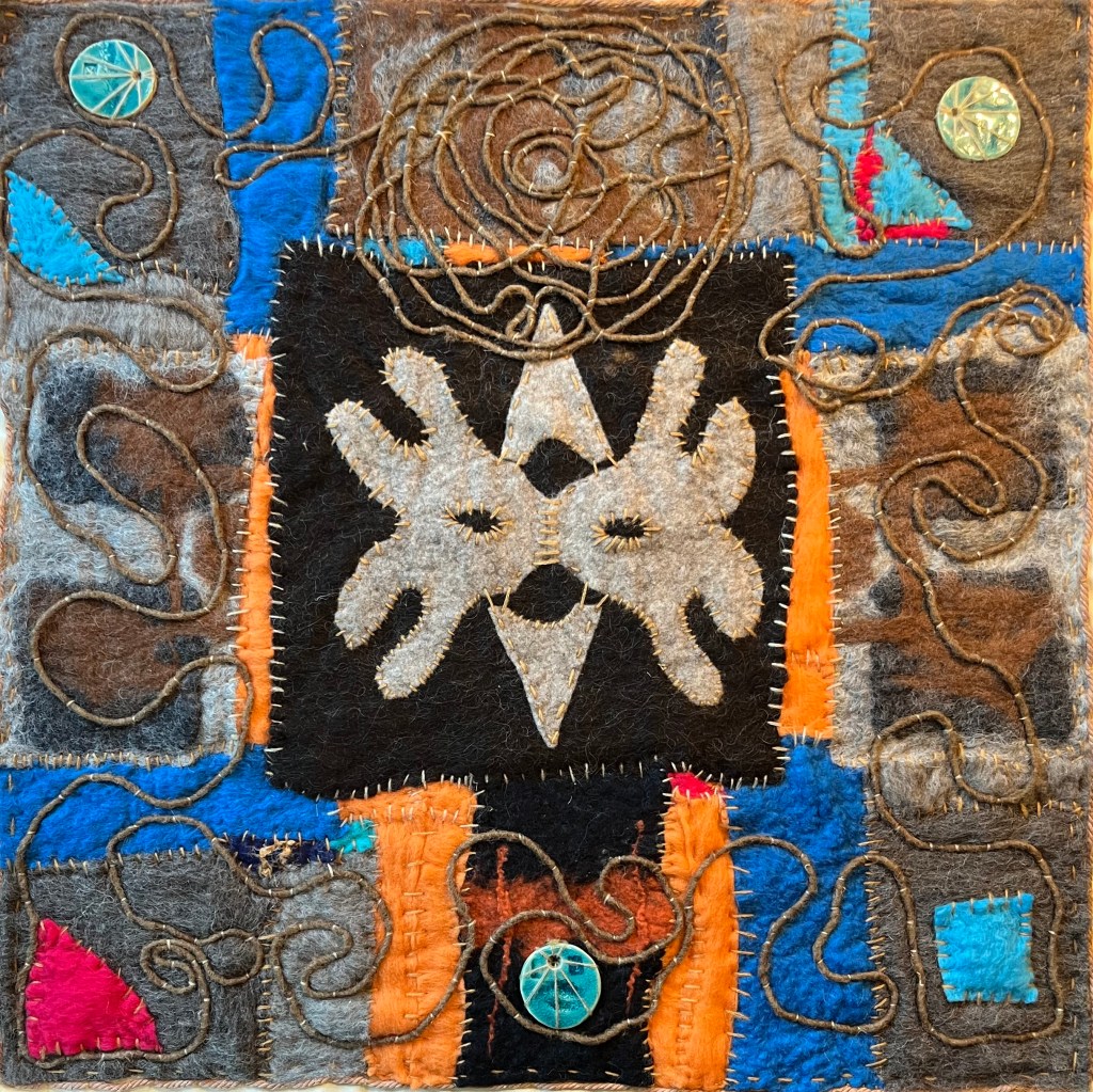 Square felt artwork with a bold symmetrical face-like motif in cream and black, surrounded by winding yarn lines, bright accents, and stitched circular emblems — blending tribal rhythm with mystical geometry.