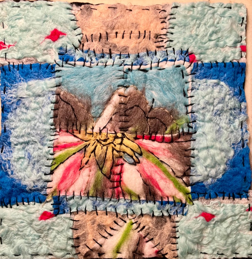 Hand-felted collage of stitched sky-blue and pink squares surrounding a central mountain range and stylized flower, combining landscape imagery with quilt-like softness.