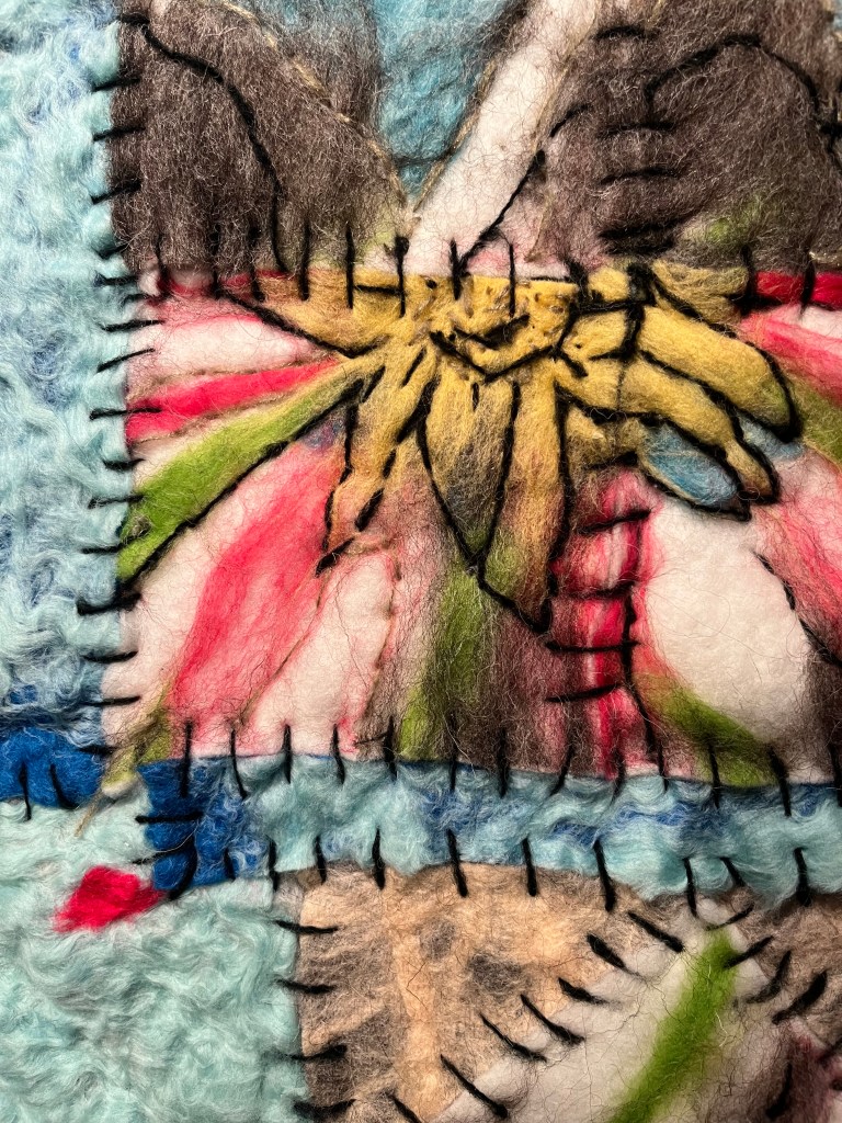Details of Hand-felted collage of stitched sky-blue and pink squares surrounding a central mountain range and stylized flower, combining landscape imagery with quilt-like softness.