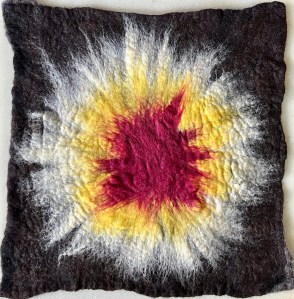 Felt textile artwork with raw, uneven edges. A central burst of deep burgundy radiates outward into rings of yellow, white, and black, forming a soft, glowing shape like a sun or unseen energy field.
